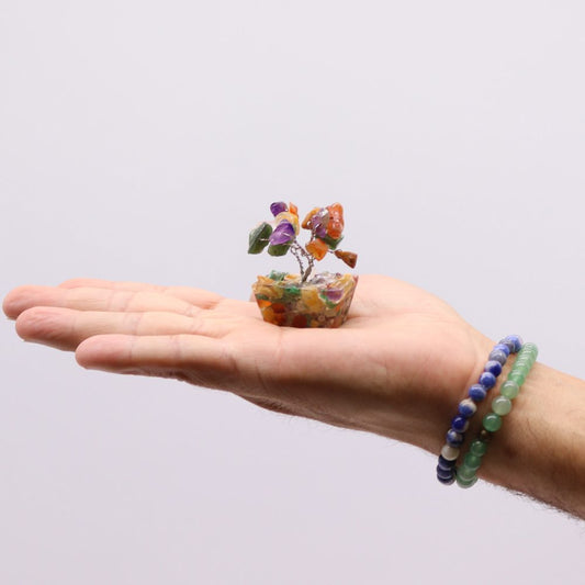 A small crystal gemstone bonsai tree sits in the palm of a hand, two beaded bracelets adorn the wrist, set against a plain white background.