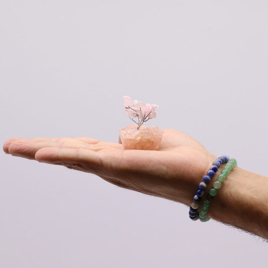 A small crystal tree sits in the center of an open palm. The wrist bears two beaded bracelets, against a plain, light background.