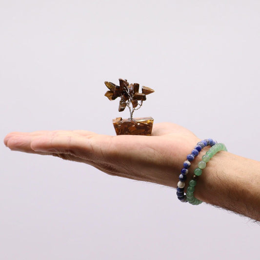 A gemstone bonsai tree rests on the open palm of a hand wearing two beaded bracelets against a plain white backdrop.