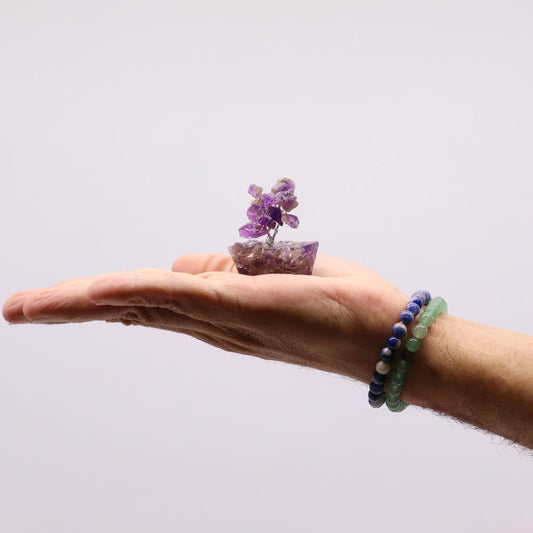 A hand holds a small, amethyst gemstone bonsai tree in its palm, presented against a plain white background, and accented by two beaded bracelets on the wrist.