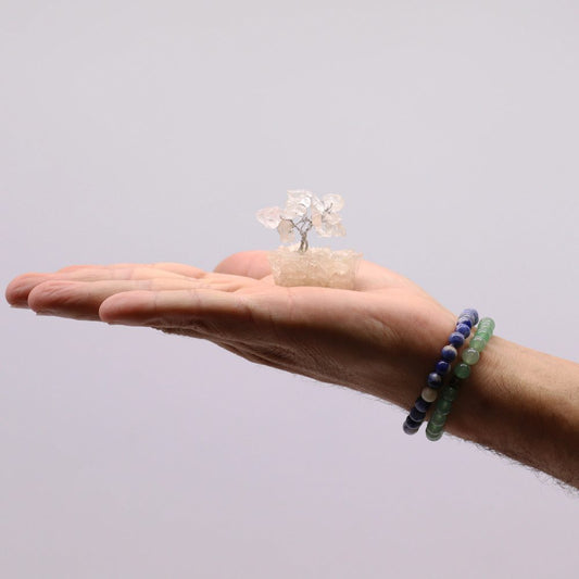 A small, quartz crystal "tree" rests in an outstretched palm, framed by a grey background. Two beaded bracelets adorn the wrist, one green and one dark blue.
