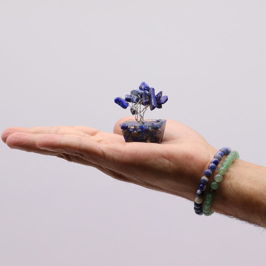 A lapis lazuli gem tree rests on the palm of a person's hand, and two beaded bracelets adorn their wrist. The background is plain white.