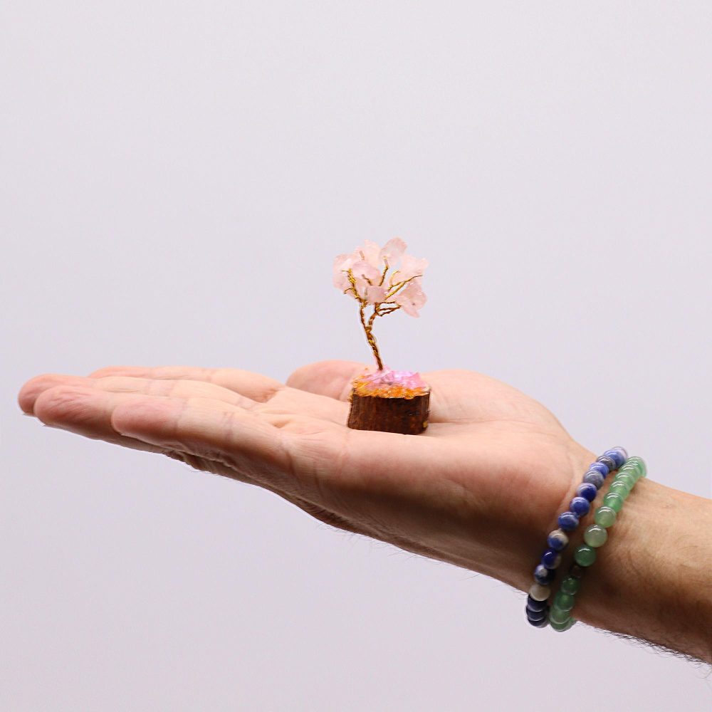 A miniature rose quartz tree, crafted from wire and stones, rests in an open palm against a white backdrop.