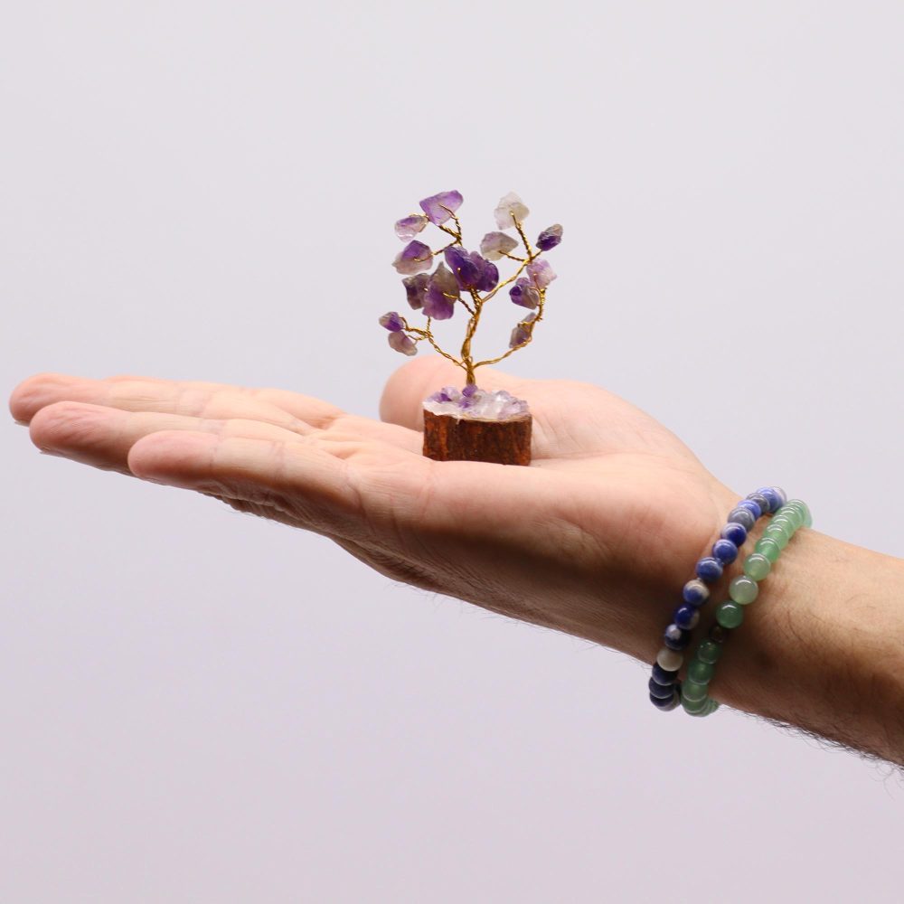 A miniature amethyst gemstone tree rests on a person’s palm. The tree is crafted from wire and small purple and clear stones. A beaded bracelet adorns the wrist.