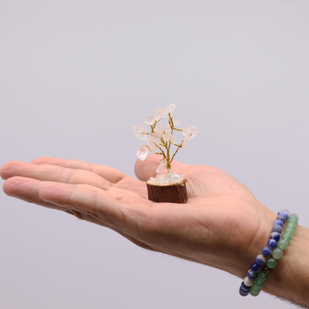 A small quartz crystal tree, crafted with gold wire, rests on a person's palm against a white background.