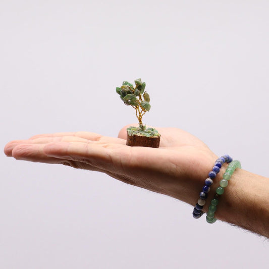 A miniature gemstone tree, crafted with wire and small green stones, rests on a person's open palm. The hand also wears two beaded bracelets; one lapis lazuli, the other green.