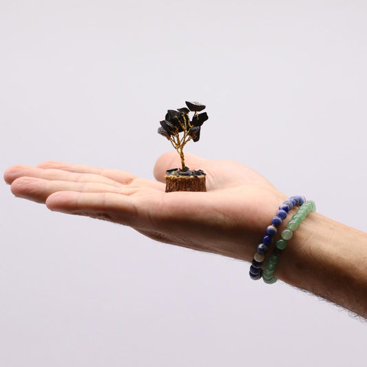 A miniature gemstone tree, crafted with wire and small black stones, rests on a person's open palm.  The hand also wears two beaded bracelets, one blue and one green.