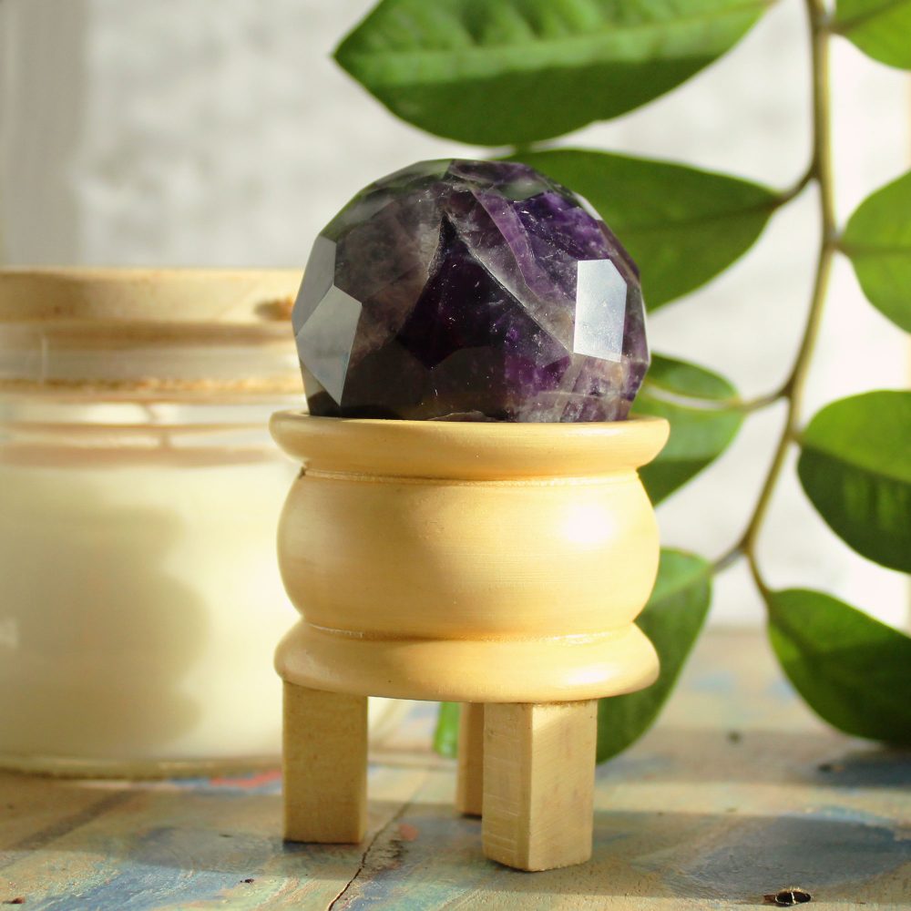 A faceted amethyst sphere rests on a small, light-colored wooden stand. The stand has three short legs. The background includes a candle and green leaves.