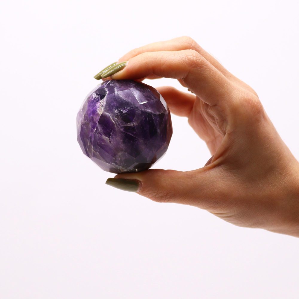 A hand holds a faceted amethyst sphere against a white background.