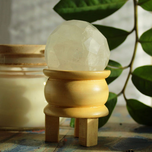 A faceted, opaque white sphere rests on a small, pale yellow wooden stand. The stand has three short legs. A candle is blurred in the background.