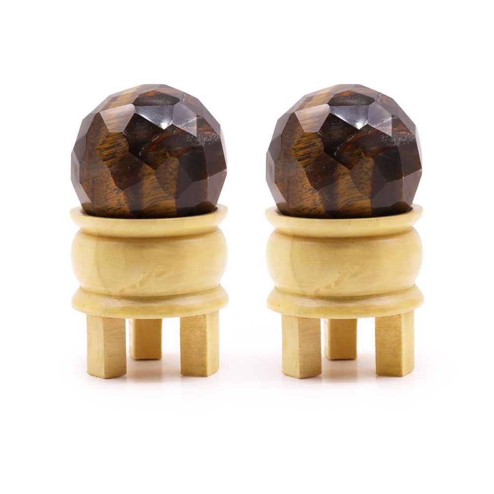 Two faceted tiger eye spheres rest in small, light-colored wooden stands with four legs, on a white background.