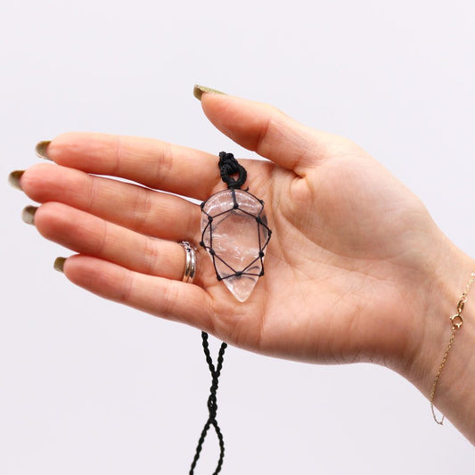 A clear crystal pendant in a black macrame holder is held in an outstretched palm against a white background.