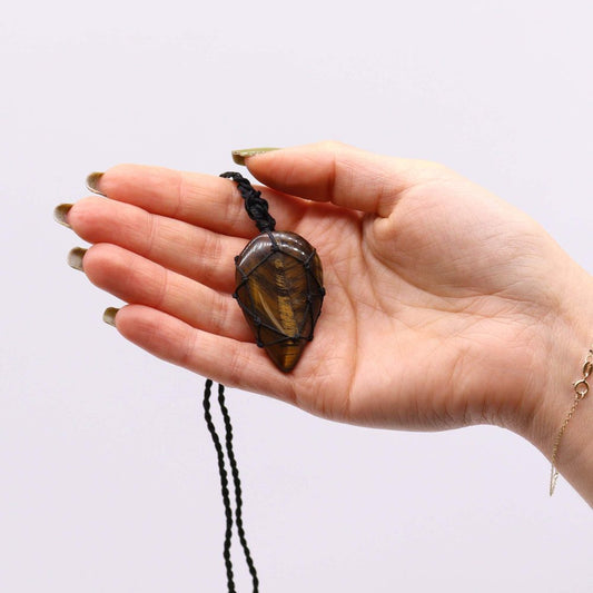 A teardrop-shaped, striated brown stone pendant hangs on a knotted black string, held in the palm of a person with a gold bracelet against a white background.