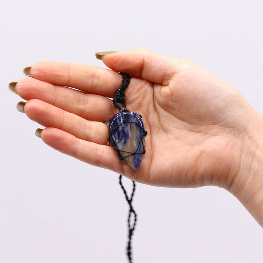 A palm holds a blue and tan crystal pendant. The crystal is wrapped in a dark cord that connects to a necklace. The background is white.