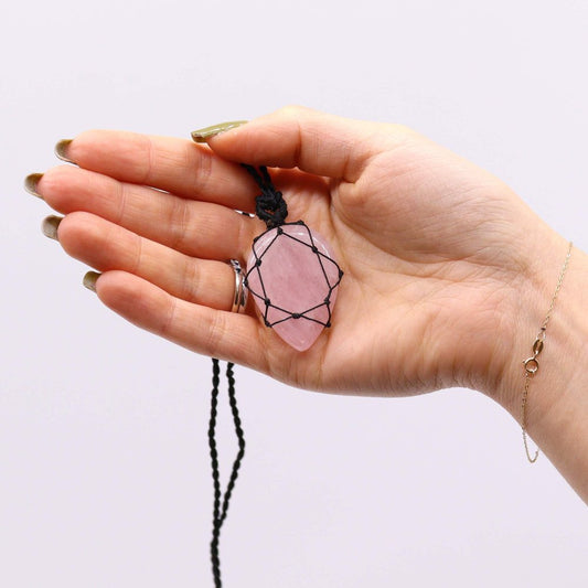 A light-skinned hand holds a pink crystal necklace with black macrame cording against a white background. A ring and gold bracelet are visible on the hand.