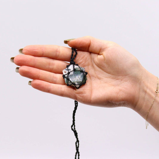 A raw crystal pendant, encased in knotted black cord, rests on an open palm. The hand is pale with manicured nails, against a plain white background.