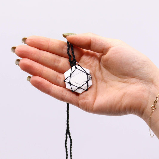 A hand holds a hexagonal white pendant with black veining, secured in a knotted black cord. The backdrop is a neutral light gray.