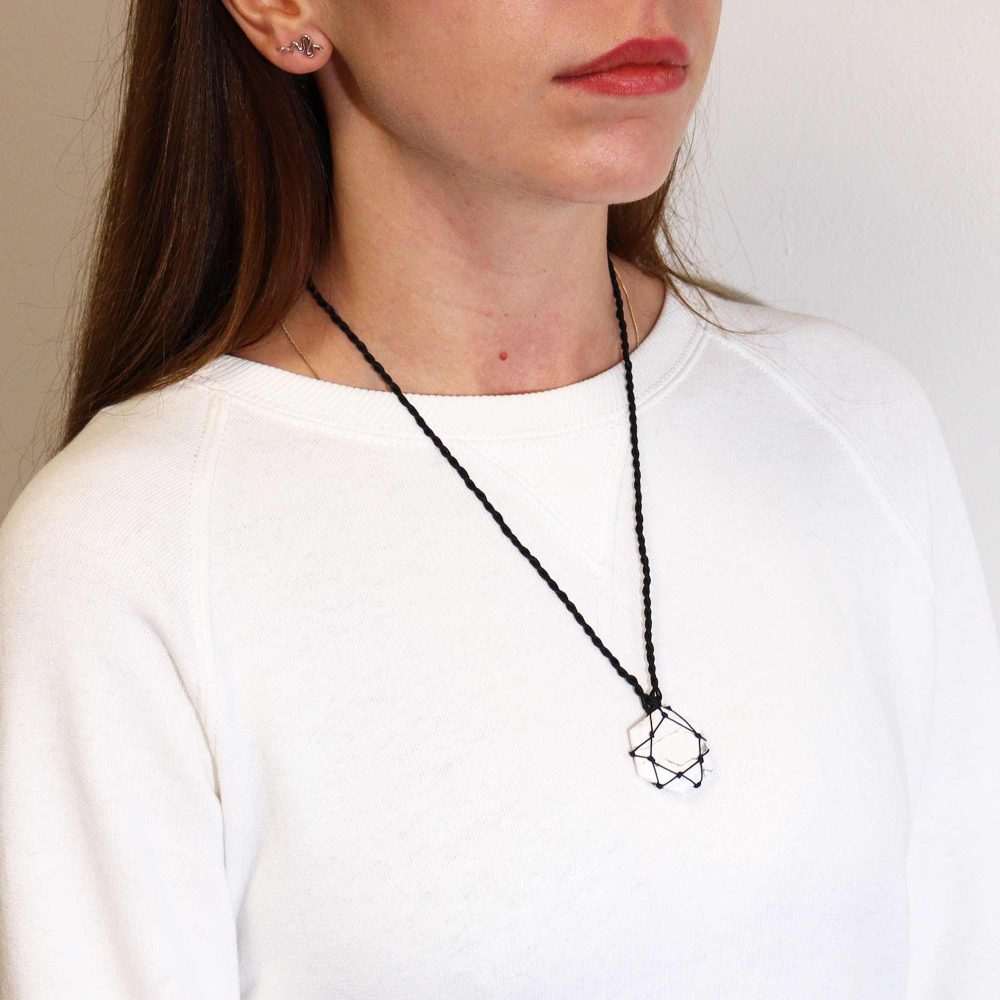 A woman wears a white geometric crystal pendant necklace, strung on a black cord. She also wears snake earrings and a white sweatshirt against a neutral wall.