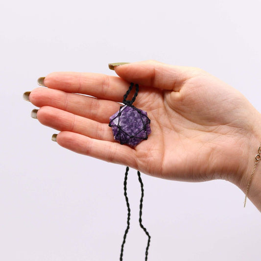 A hand holds a purple, star-shaped crystal pendant wrapped in black cord, presented against a plain white background. A delicate gold bracelet is visible on the wrist.