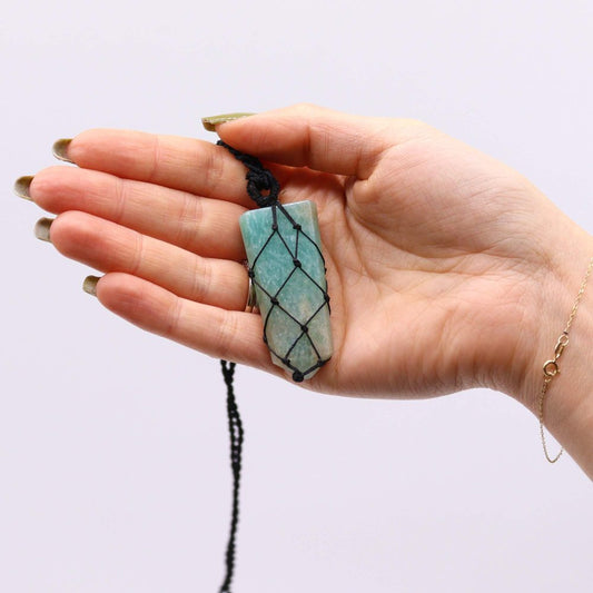 A person's open hand holds a crystal pendant, wrapped in black macrame cord and suspended from a black string, against a white background.