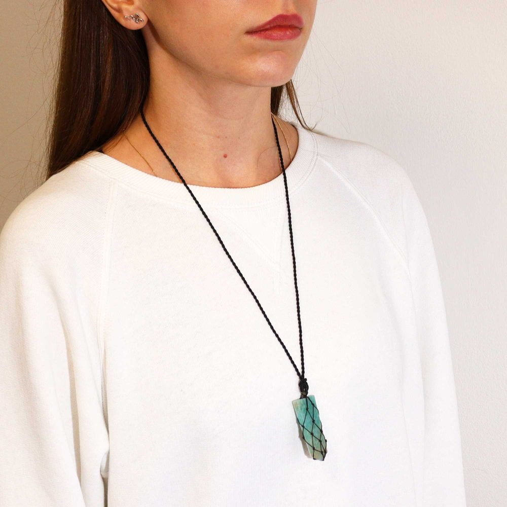 A woman wears a rectangular, blue-green stone pendant hanging from a black cord necklace. She is in front of a plain, white wall and wears a white sweater.