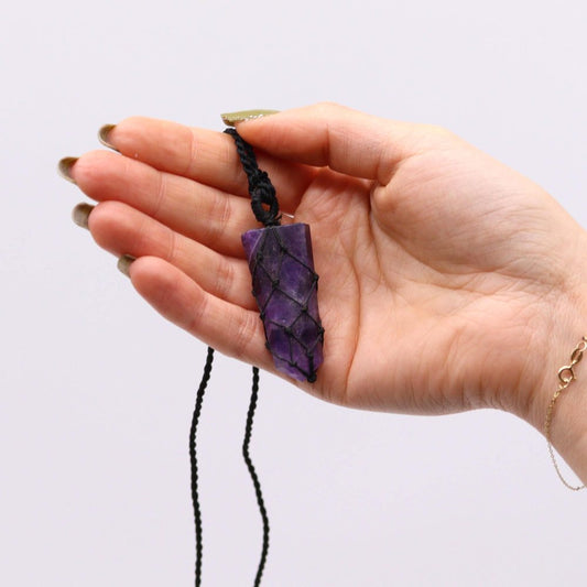 A hand holds an amethyst crystal pendant. The purple crystal is wrapped in black knotted cording, hanging against a white background.