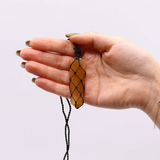 A hand is holding a vertically oriented, orange crystal pendant that is encased in black macrame. The background is white.
