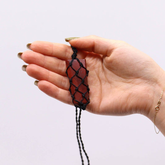 A hand holds a rectangular, reddish-brown crystal encased in a black macrame net. The background is a plain white, and a delicate gold bracelet adorns the wrist.