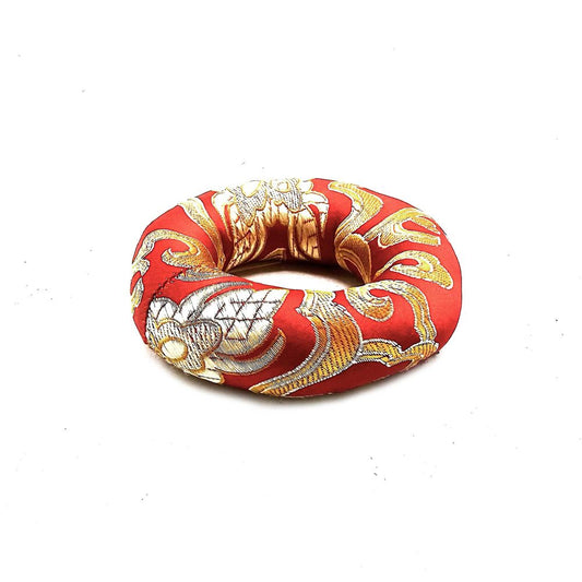 A red and gold ring-shaped cushion rests on a white surface. The cushion is decorated with an ornate embroidered pattern.