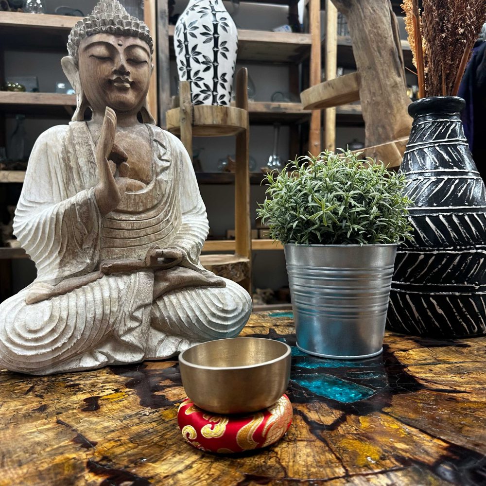 A brass singing bowl rests on a red cushion, sitting on a wooden table near a wooden Buddha statue and potted plant.