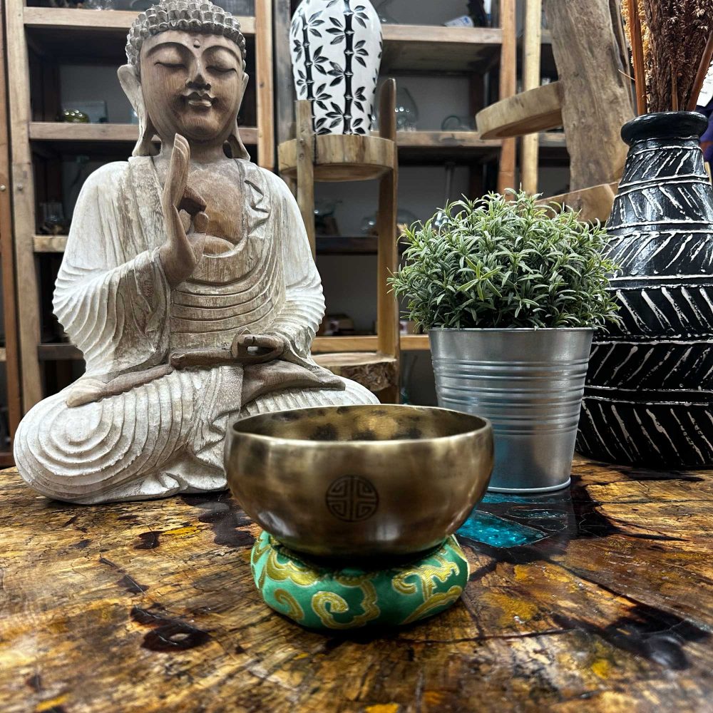 A brass singing bowl rests on a teal cushion. It sits on a wooden table beside a wooden Buddha statue. A small potted plant is nearby.