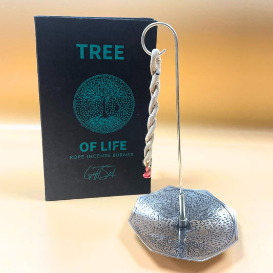 A metal incense holder stands with a lit rope incense hanging. Behind it, a black box reads "TREE OF LIFE ROPE INCENSE BURNER Gift Set," depicting a tree design. The background is tan.