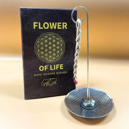 An incense burner holds a twisted rope, standing next to a brown package labeled "FLOWER OF LIFE ROPE INCENSE BURNER Gift Set," featuring a geometric design. The background is plain beige.