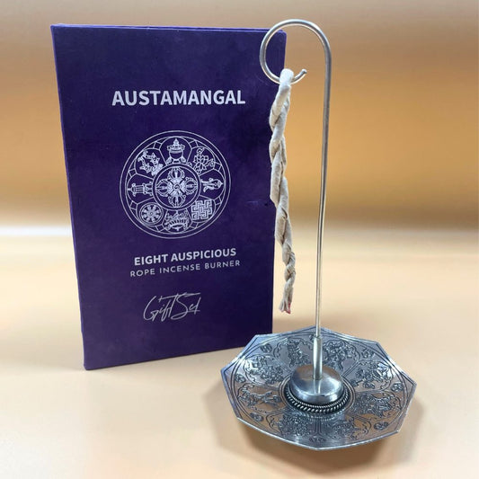 A decorative rope incense burner with a twisted rope hangs on a metal stand over a detailed octagonal base, beside a purple box labeled "AUSTAMANGAL EIGHT AUSPICIOUS ROPE INCENSE BURNER Gift Set."