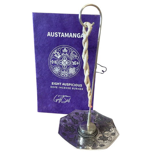 An intricate metal incense holder supports a twisted rope incense, placed before a purple box labeled "AUSTAMANGA: EIGHT AUSPICIOUS ROPE INCENSE BURNER" with symbolic designs on its surface.