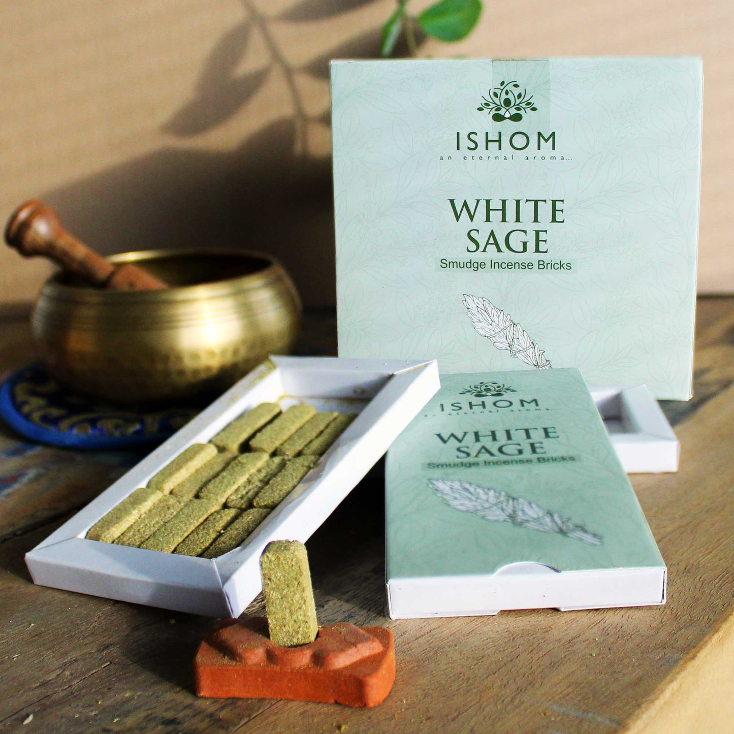 A box labeled "ISHOM WHITE SAGE Smudge Incense Bricks" is open, revealing rectangular incense bricks. A single brick sits on a terracotta holder beside a brass bowl.