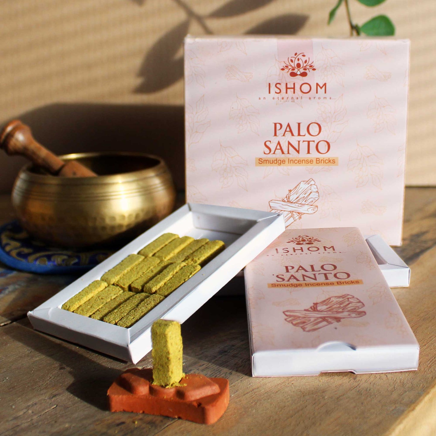 Incense bricks displayed in an open box titled "ISHOM Palo Santo Smudge Incense Bricks." A single brick stands upright on a clay holder. A brass bowl is in the background.