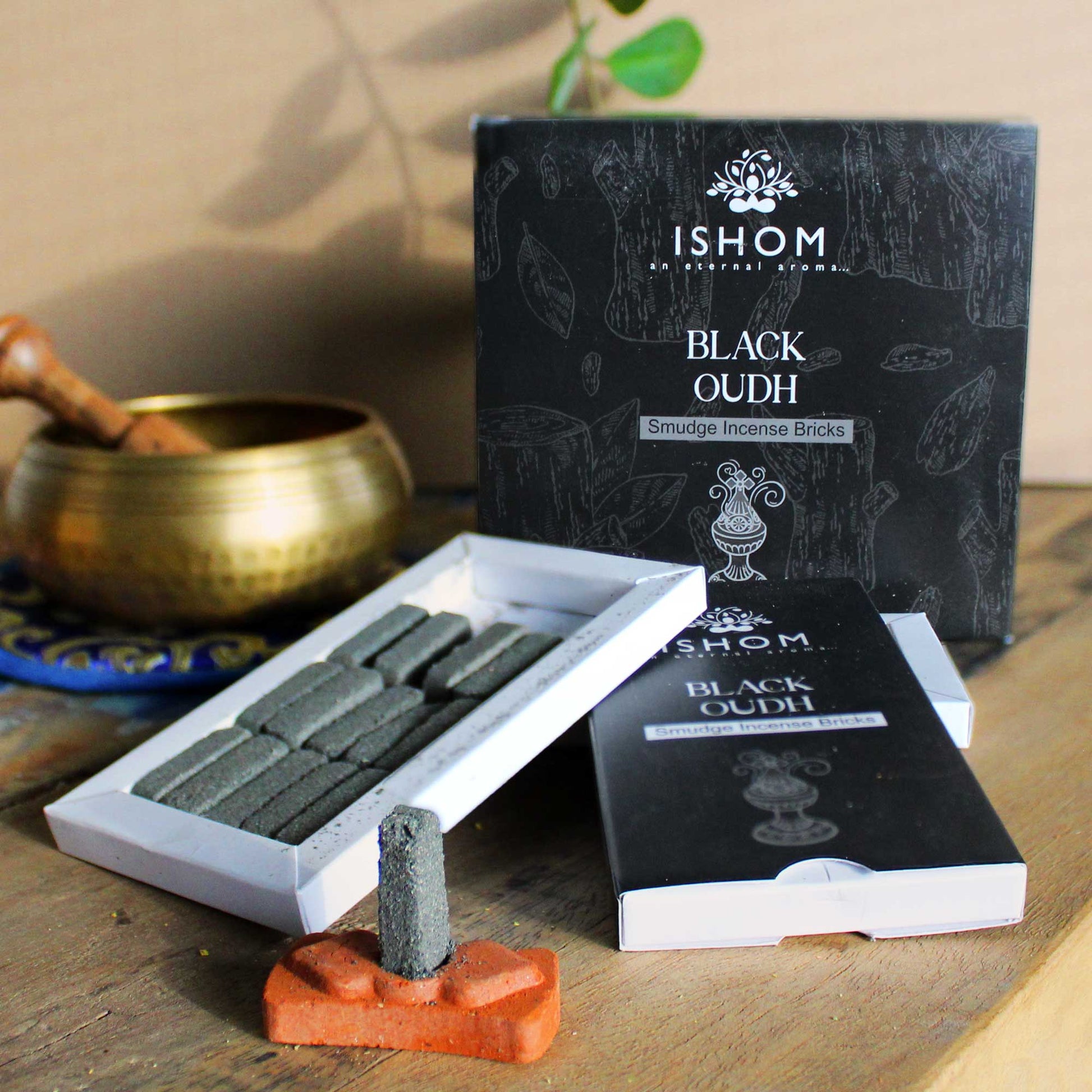 Incense bricks and packaging labeled "ISHOM BLACK OUDH Smudge Incense Bricks" are placed on a wooden surface. A gold bowl and a leafy plant are visible in the background.