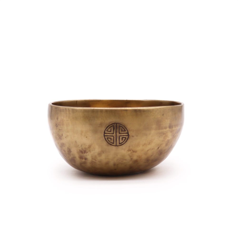 A brass singing bowl sits on a plain background, featuring an engraved circular design on its side, reflecting light with its smooth, polished surface.