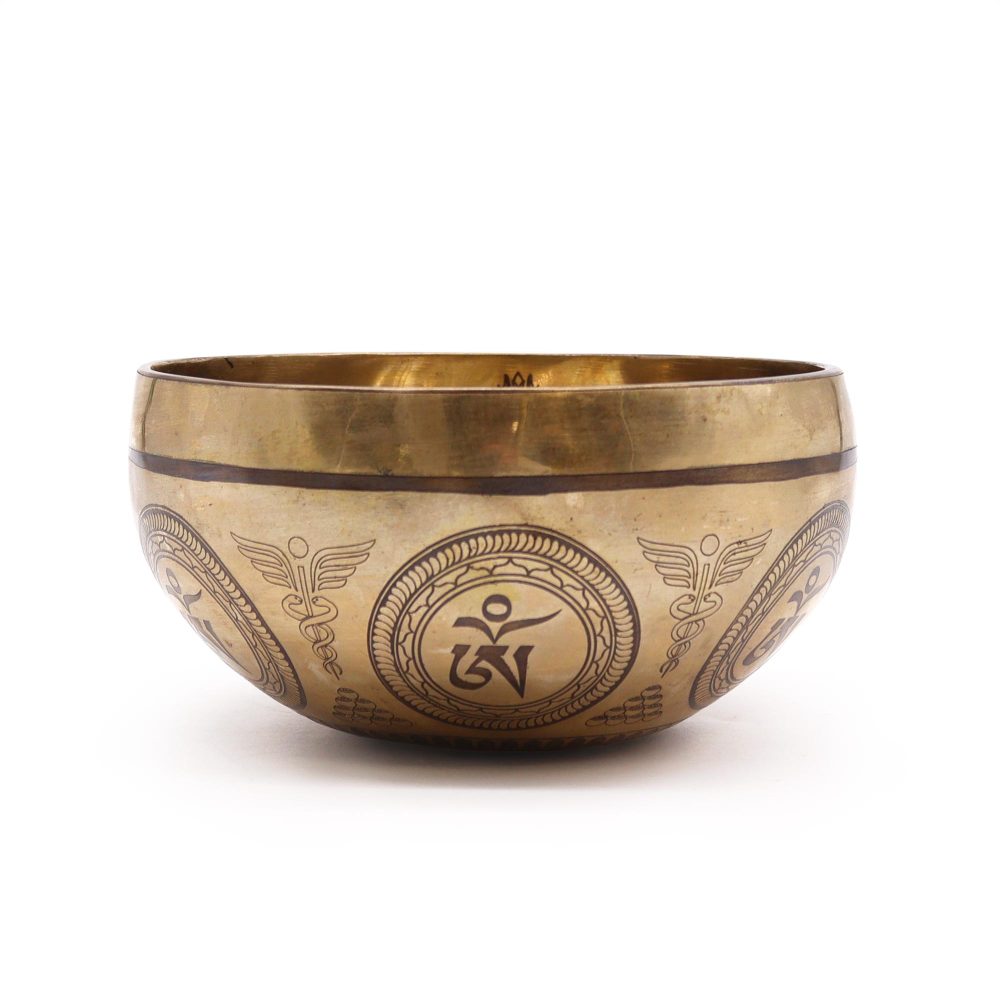 A bronze singing bowl is resting on a flat surface, engraved with intricate patterns and symbols, including "Om" characters and decorative motifs, in a bright, minimalist setting.