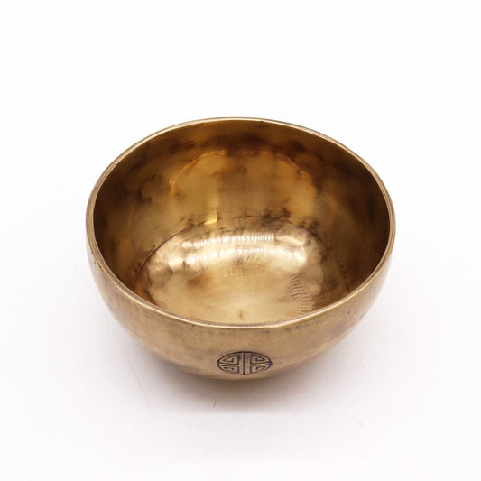 A brass singing bowl sits upright on a plain white background, featuring a circular symbol on its outer surface, reflecting light across its polished interior.