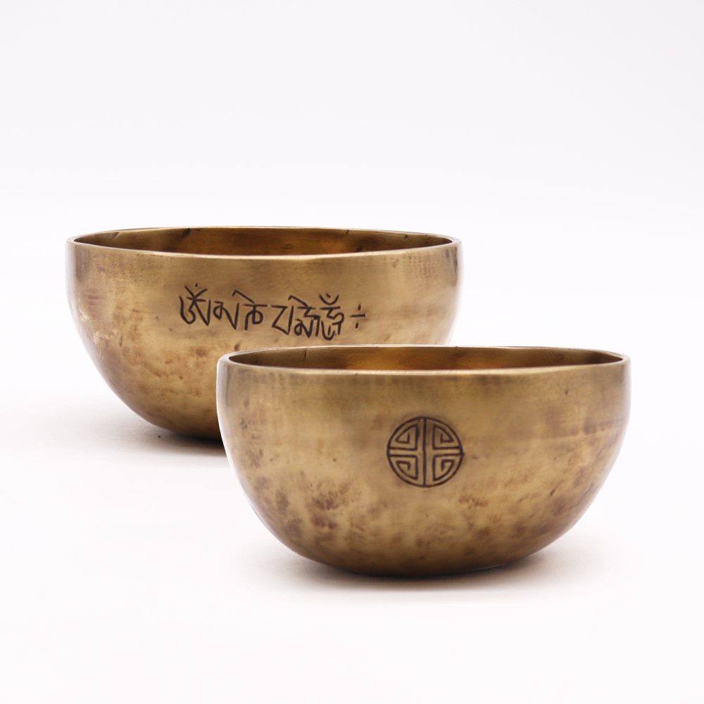 Two brass bowls with ornate engravings sit on a white background. The rear bowl features text: "ॐ मणि पद्मे हूं". The front bowl has a circular geometric design.