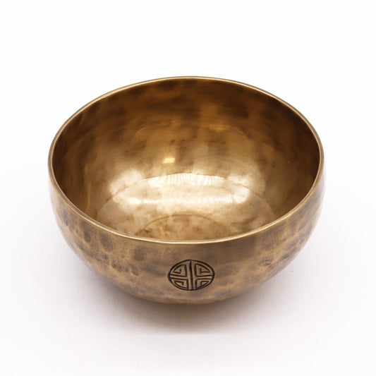 A brass singing bowl sits quietly on a plain white surface. It has a hammered texture with a circular symbol etched on the side, reflecting light subtly.