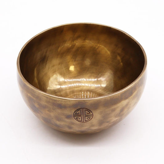 A bronze bowl sits on a white surface, featuring a textured finish and an engraved circular symbol on its exterior. The bowl's interior is smooth and reflective.