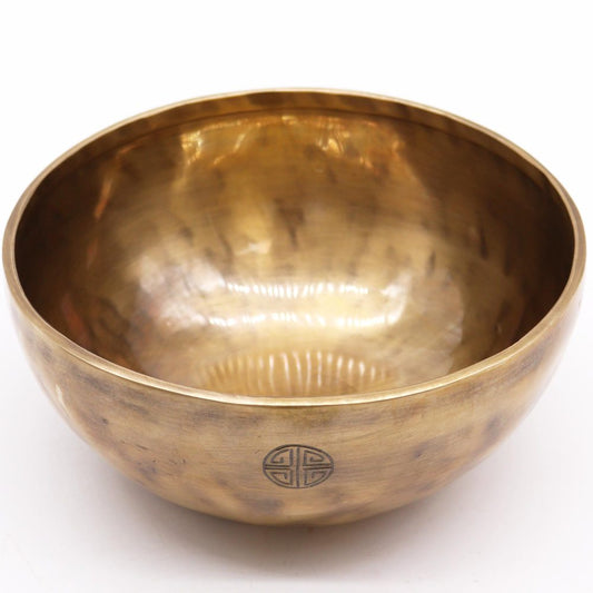 A polished bronze singing bowl sits on a plain surface, featuring a circular symbol on its side. The interior gleams with light reflections, suggesting a well-crafted, smooth finish.