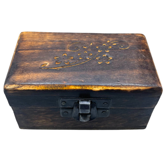 A dark wooden box with an intricate floral carving on the lid sits closed, fastened by a black metal latch, against a plain white background.