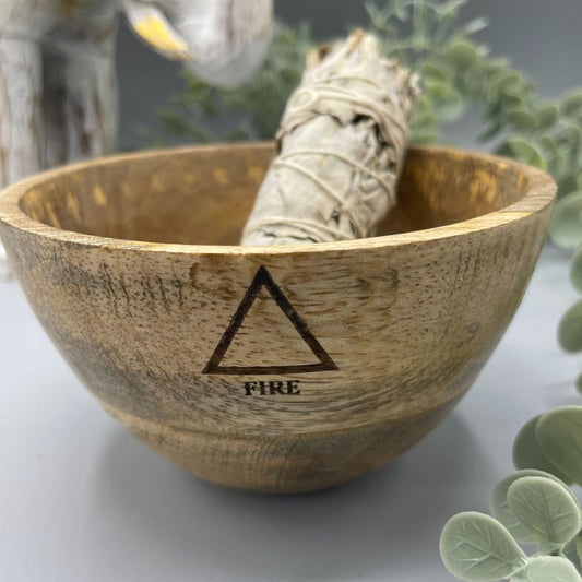 Sage bundle sits in a wooden bowl, which is etched with an upward-pointing triangle and the word “FIRE.” The bowl is set on a gray surface with other decorative elements in the background.