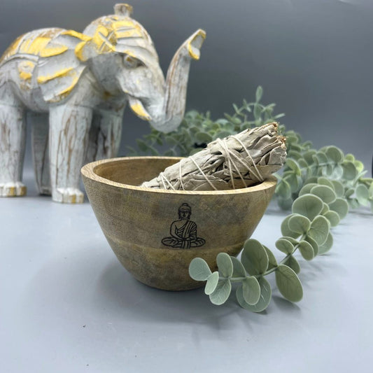 A bundle of white sage tied with string sits in a wooden bowl that has a drawing of a Buddha on its side, placed on a gray surface, with a white and gold elephant figure in the background.