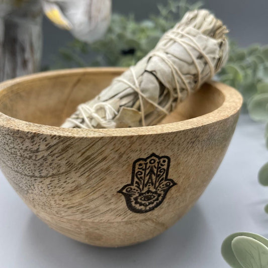 A sage bundle rests in a wooden bowl, which is decorated with a hamsa illustration. A neutral background with plants is subtly visible.