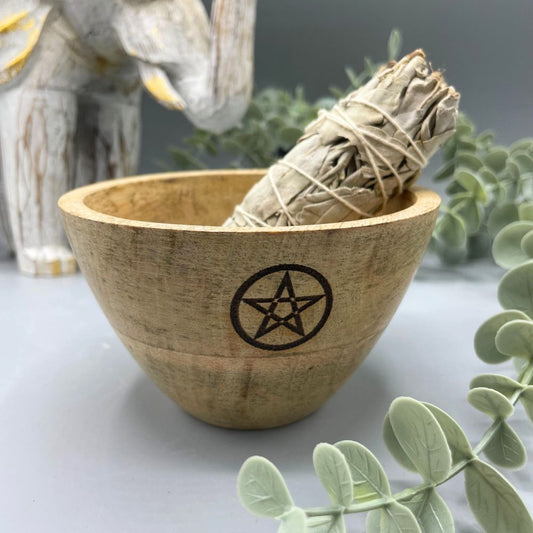 A sage bundle rests in a wooden bowl with a pentagram symbol. The bowl sits on a gray surface with decorative greenery and an elephant statuette in the background.
